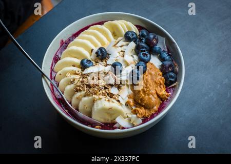 Bol à smoothie avec de la poudre d'açaï, des bleuets, du beurre d'arachide, de la banane, des flocons de noix de coco rôtis et du granola. Banque D'Images