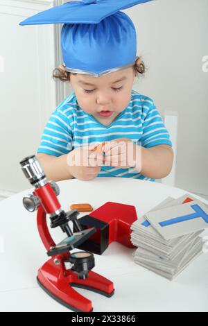 Petit garçon en chapeau bleu de graduation assis à table avec microscope rouge Banque D'Images