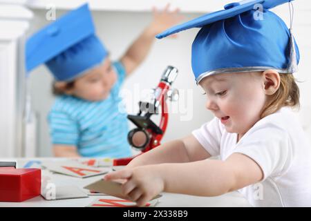 Heureuse petite fille dans le chapeau bleu de graduation dispose des cartes avec des lettres sur la table à côté du garçon avec microscope Banque D'Images