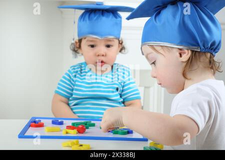 Heureuse petite fille et garçon sérieux dans le chapeau bleu de graduation assis à table et jouant avec le tableau magnétique des enfants avec des lettres colorées Banque D'Images