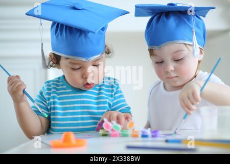 Petite fille et garçon dans les chapeaux bleus de graduation peignent des couleurs à table Banque D'Images