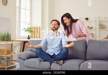 La femme en colère se querelle et crie à l'homme qui est assis en position méditative sur le canapé à la maison. Banque D'Images