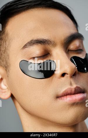 Bel homme asiatique subissant une routine beauté et soin de la peau, portant des patchs noirs pour les yeux dans un cadre de studio gris. Banque D'Images