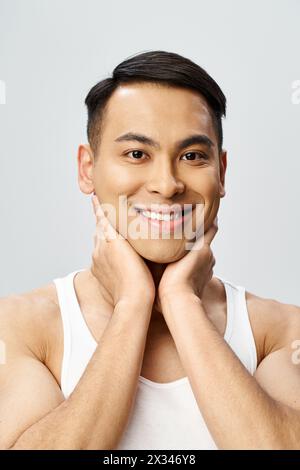 Un bel homme asiatique avec un sourire joyeux, les mains reposant sur son menton dans un décor gris de studio. Banque D'Images