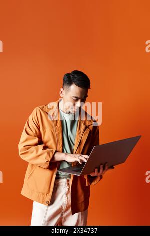 Un homme asiatique élégant dans une veste orange engrossé dans le travail sur un ordinateur portable dans un cadre de studio. Banque D'Images