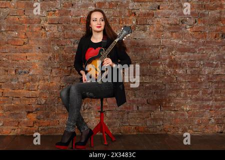 Jolie jeune femme avec mini guitare est assise sur le tabouret dans le studio avec le mur de briques rouges Banque D'Images