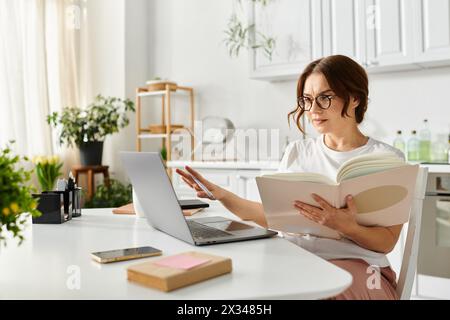 Femme d'âge moyen englouti dans un livre, multitâche avec un ordinateur portable. Banque D'Images