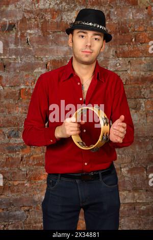 Bel homme en chapeau pose avec tambourin dans le studio avec mur de briques Banque D'Images