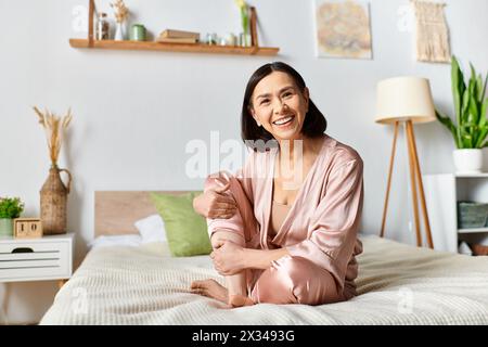 Une femme mature dans des vêtements pour la maison confortables est assise au sommet d'un lit, souriant avec satisfaction. Banque D'Images