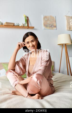 Une femme mature dans des vêtements de maison confortables est assise sur un lit, souriante. Banque D'Images