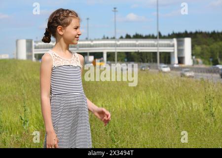 Jolie fille se tient sur l'herbe verte et regarde loin près de la route le jour d'été Banque D'Images