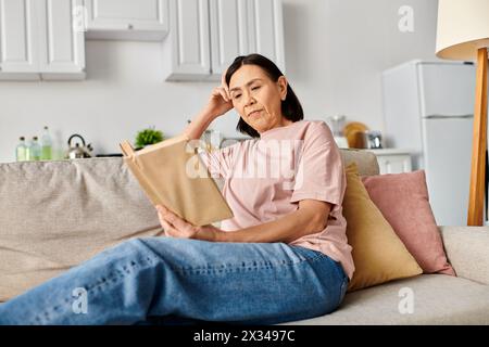 Une femme mature dans des vêtements confortables est assise sur un canapé, engrossée dans la lecture d'un livre. Banque D'Images