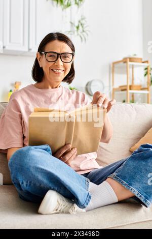 Femme mature dans des vêtements confortables assis sur le canapé, engrossée dans la lecture d'un livre. Banque D'Images