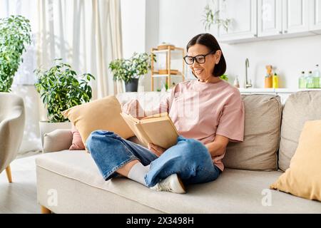 Femme mature dans une tenue confortable engrossée dans un livre tout en étant assise sur un canapé confortable. Banque D'Images