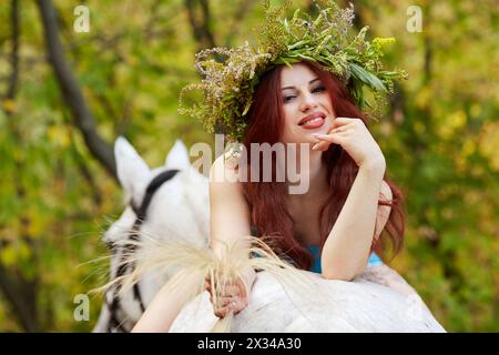 Jeune femme rousse souriante avec couronne florale sur la tête se trouve à cheval dans le parc. Banque D'Images