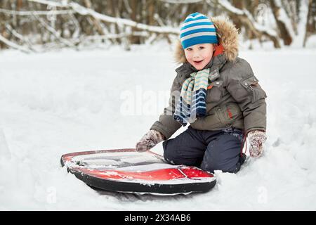 Petit garçon est assis sur la neige près de traîneaux souples dans le parc d'hiver. Banque D'Images