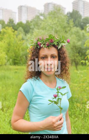 portrait demi-longueur de jeune femme avec couronne de fleurs zone boisée urbaine près du complexe d'appartements, tenant le trèfle, souriant Banque D'Images