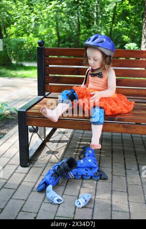 Petite fille s'assoit sur le banc et enlève les patins à roulettes dans le parc vert d'été Banque D'Images