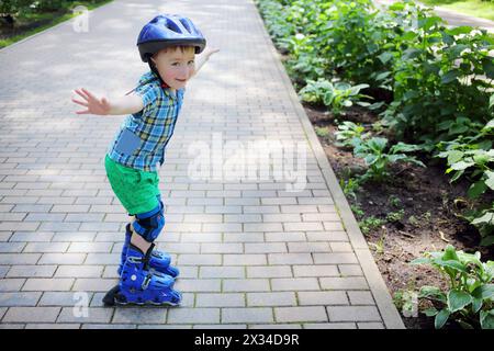 Petit garçon dans des patins à roulettes de casque comme oiseau dans le jardin le jour d'été Banque D'Images