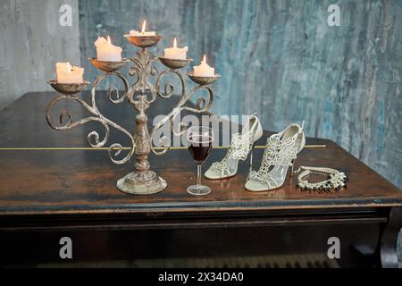 Vieux piano à queue noir avec des bougies allumées en chandelier, verre de vin rouge, chaussures à talons hauts et collier sur le couvercle dans la chambre avec des murs déchiquetés. Banque D'Images