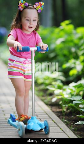 Petite fille mignonne en short ride scooter dans le jardin ensoleillé d'été Banque D'Images