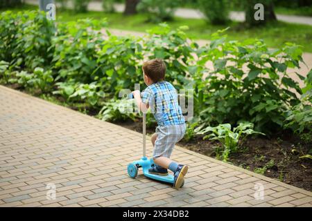 Petit garçon en short monte scooter dans le parc vert d'été Banque D'Images