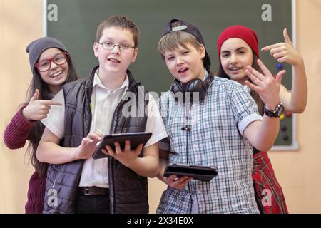 deux garçons et deux filles avec des écouteurs et une tablette posant pour la caméra sur fond de tableau noir vert dans la salle de classe, les filles montrent le geste et le sourire, focu Banque D'Images