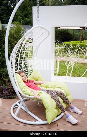 Petite fille souriante est assise dans la balançoire blanche accrochée à la chaîne dans le parc d'été. Banque D'Images