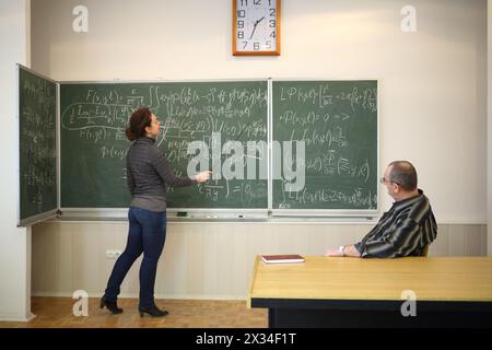 La femme parle avec un pointeur près du tableau noir avec des formules et l'homme l'écoute dans la salle de classe de l'école Banque D'Images