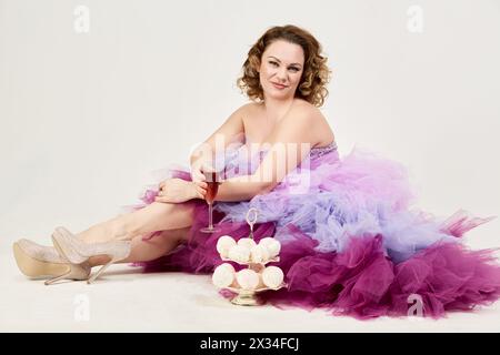 Femme souriante dans une robe mauve moelleuse se trouve sur le sol près d'un plat de deux étages avec des guimauves tenant un verre de vin à la main. Banque D'Images