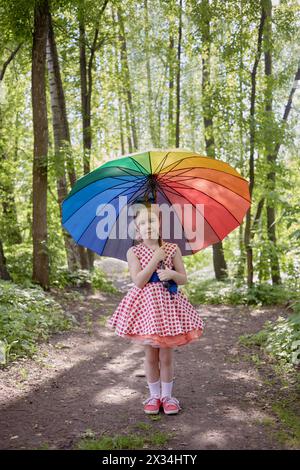 Petite fille en robe à pois se dresse avec parapluie couleur arc-en-ciel dans le parc d'été. Banque D'Images