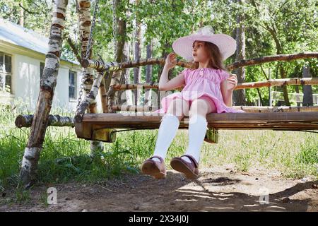 Petite fille en robe rose et chapeau de soleil se trouve sur des balançoires en bois sous les arbres. Banque D'Images