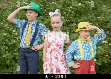 Deux garçons en chapeaux et une fille se tiennent dehors contre des buissons verts. Banque D'Images