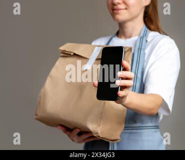 Serveuse dans un tablier tenant un sac en papier avec de la nourriture à emporter et montrant l'écran du smartphone. Paiement mobile avec qr-code, commande alimentaire en ligne. Banque D'Images