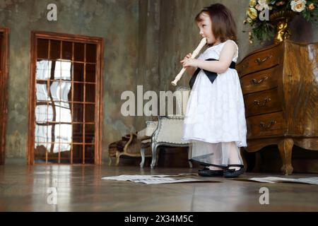 Petite fille en robe blanche joue de la flûte dans la chambre avec des notes sur le sol Banque D'Images