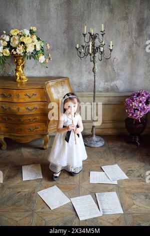 Petite fille en robe blanche joue de la flûte dans la chambre avec des notes sur le sol Banque D'Images