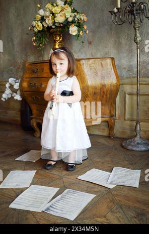 Jolie petite fille en robe blanche joue de la flûte dans la chambre avec des notes au sol Banque D'Images