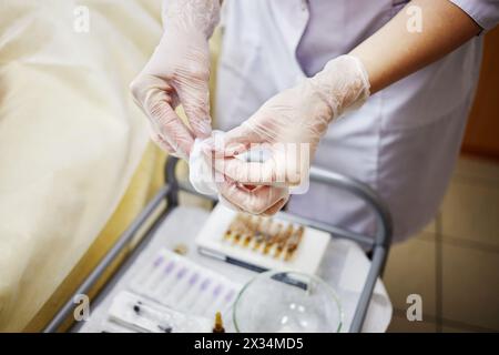 Les mains féminines dans des gants en caoutchouc tiennent le coton sur la table médicale avec des ampoules et des seringues. Banque D'Images