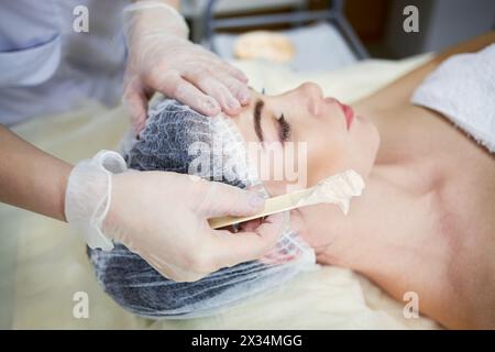 Mains de l'esthéticienne appliquer masque cosmétique avec spatule en bois sur le visage de la femme couchée sur le canapé dans le salon de beauté. Banque D'Images