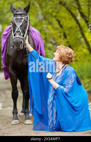 Femme souriante aux cheveux rouges en bleu capote avec cheval de baie dans le parc. Banque D'Images