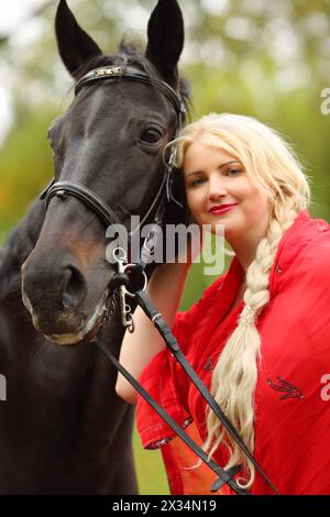 Femme en robe rouge et cheveux blonds se tient près du cheval dans le parc Banque D'Images