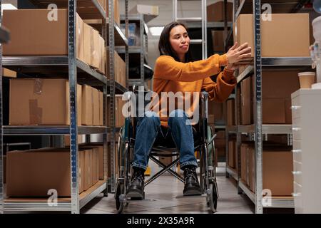 Femme utilisateur de fauteuil roulant prenant la boîte en carton de l'étagère pour préparer la commande du client pour l'expédition dans l'entrepôt. Manipulateur de colis asiatique tenant le colis, travaillant dans un entrepôt favorable aux personnes handicapées Banque D'Images