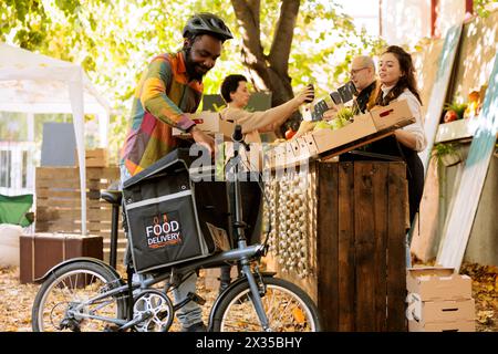 Courrier masculin noir mettant la boîte de produits biologiques dans le sac à dos pour livrer la commande aux clients, en utilisant le vélo. Jeune livreur joyeux travaillant sur l'expédition des légumes du marché des agriculteurs. Banque D'Images