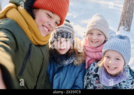 L'un des quatre enfants heureux prenant selfie sur smartphone tout en se tenant à côté de ses amis joyeux portant des vêtements d'hiver chauds Banque D'Images