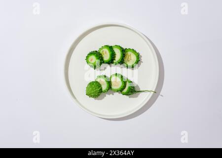 Sur un fond blanc et un plat rond en céramique contenant des tranches fraîches de melon amer. Scène publicitaire. La gourde amère est connue comme un effet simple et Banque D'Images