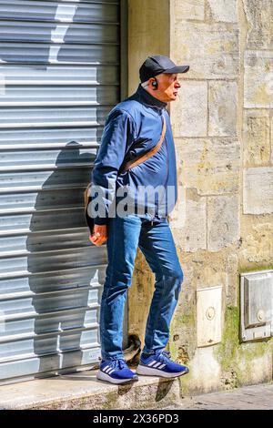 Homme plus âgé vêtu de façon décontractée debout devant des volets fermés en attendant l'ouverture du magasin - Tours, Indre-et-Loire (37), France. Banque D'Images