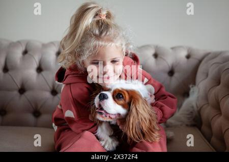 Belle fille blonde enfant est assis à la maison sur un canapé, jouant avec et embrassant le chien, créant des moments de joie et de compagnie avec l'animal de compagnie. Banque D'Images