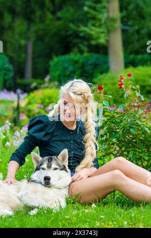 Jeune femme blonde caresse chien Husky assis sur l'herbe dans le parc d'été. Banque D'Images