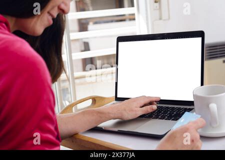 vue arrière d'une femme adulte latine heureuse assise sur le lit dans sa chambre à coucher payant pour internet écrire les détails de la carte sur ordinateur portable avec écran blanc, espace de copie. Banque D'Images