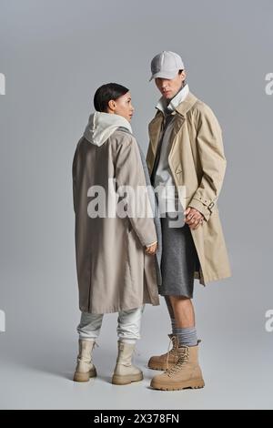 Un jeune couple élégant en trench-coat debout l'un à côté de l'autre dans un studio sur un fond gris. Banque D'Images